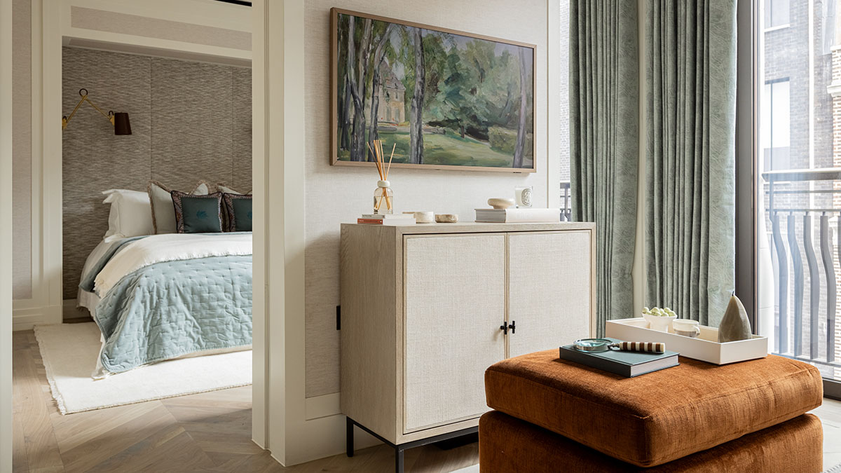 Furnished bedroom and living area with a neutral cabinet, rust-coloured ottoman and soft blue bedding for a move-in-ready home.